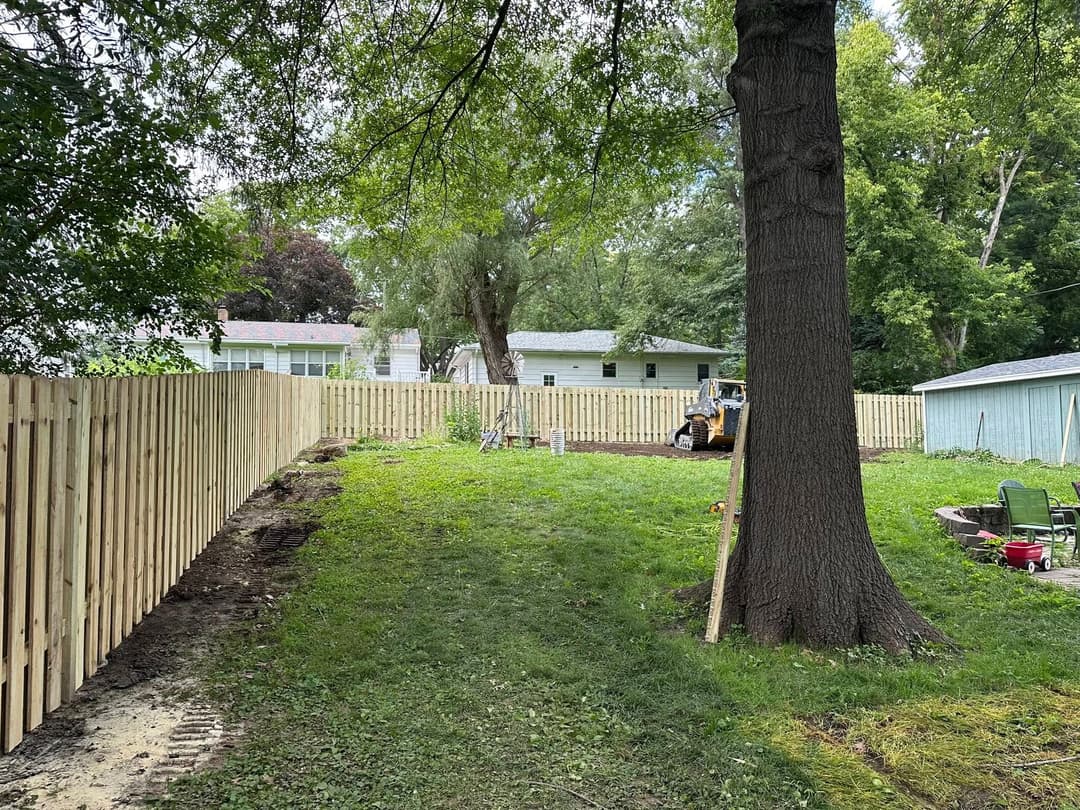 Newly installed wooden fence surrounding a green lawn with a large tree and nearby buildings.