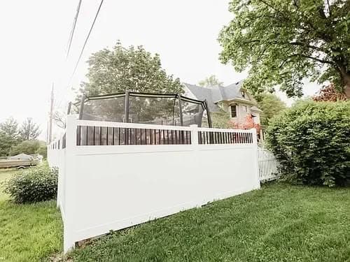 Backyard with a trampoline enclosed by a white fence and lush greenery.