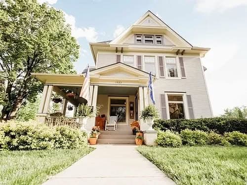 Charming historic house with a welcoming porch, surrounded by green landscaping and flowers.