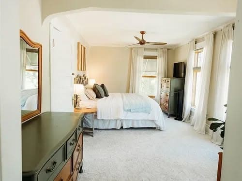 Cozy bedroom with light colors, a ceiling fan, and natural light from large windows.