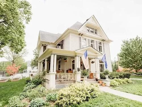 Victorian-style house with flags, surrounded by lush greenery and flowers. Perfect for historic architecture lovers.