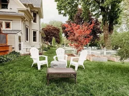 Outdoor garden seating area with white chairs, a table, and colorful trees near a house.