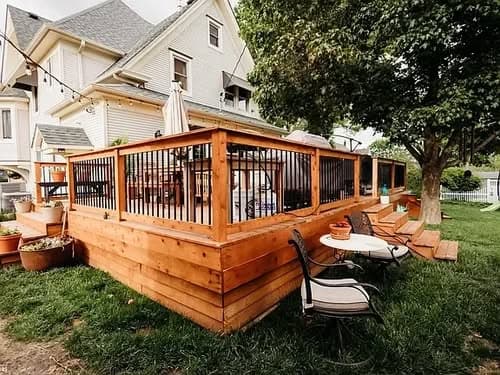 Wooden deck with black railings and seating, surrounded by greenery near a large house.