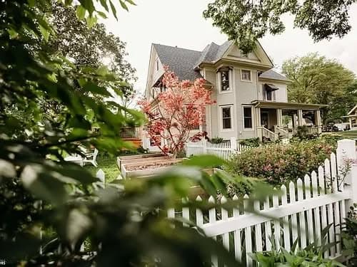 Victorian-style home with a colorful tree and white picket fence surrounded by greenery.
