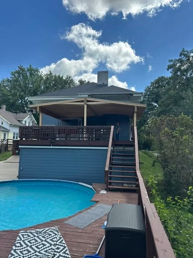 Modern backyard with a swimming pool, wooden deck, and blue house under a partly cloudy sky.