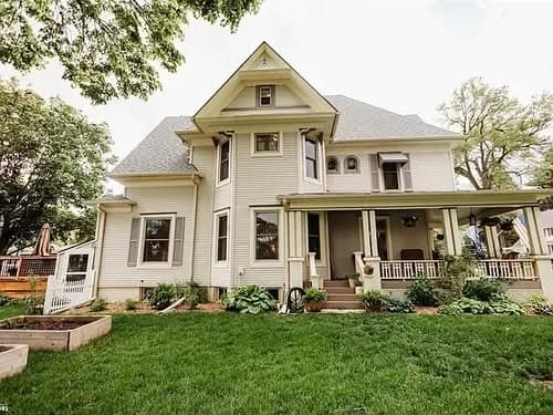 Victorian-style house with porch, landscaped yard, and large windows under a cloudy sky.