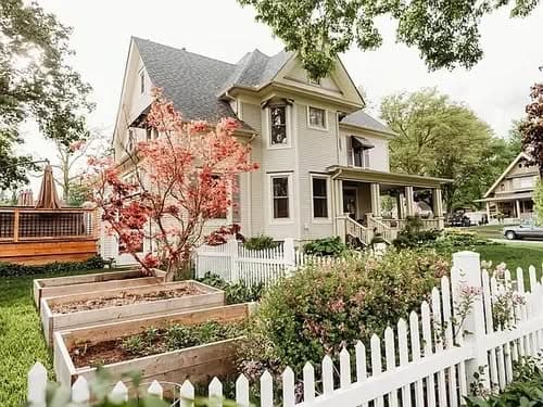 Victorian-style house with garden beds, white fence, and blooming trees in a vibrant neighborhood.