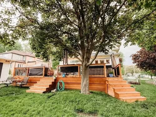 Wooden deck with steps under a large tree, featuring a house in the background and a green lawn.