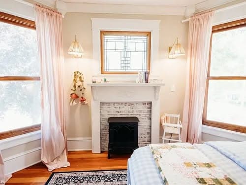 Cozy bedroom featuring a vintage fireplace, soft pink curtains, and natural light.