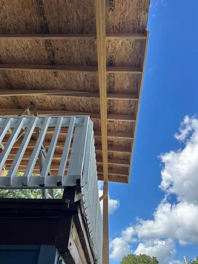 View of a wooden roof structure over a porch under a bright blue sky with clouds.