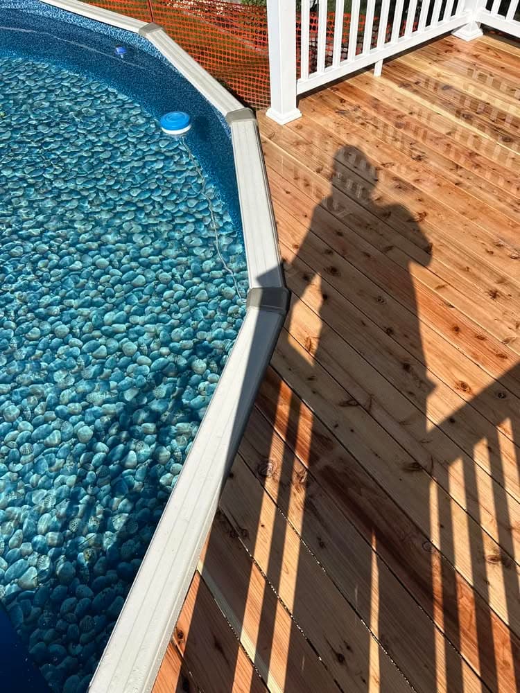 Shadow of a person by a swimming pool with a pebble bottom and wooden deck.