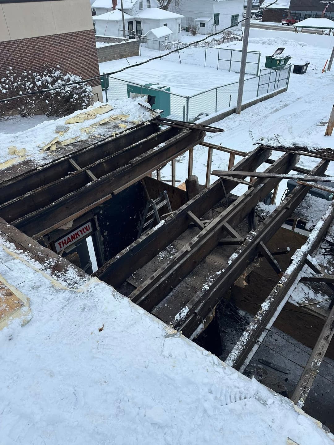 Damaged building roof with visible wooden beams, snow-covered ground, and construction site.