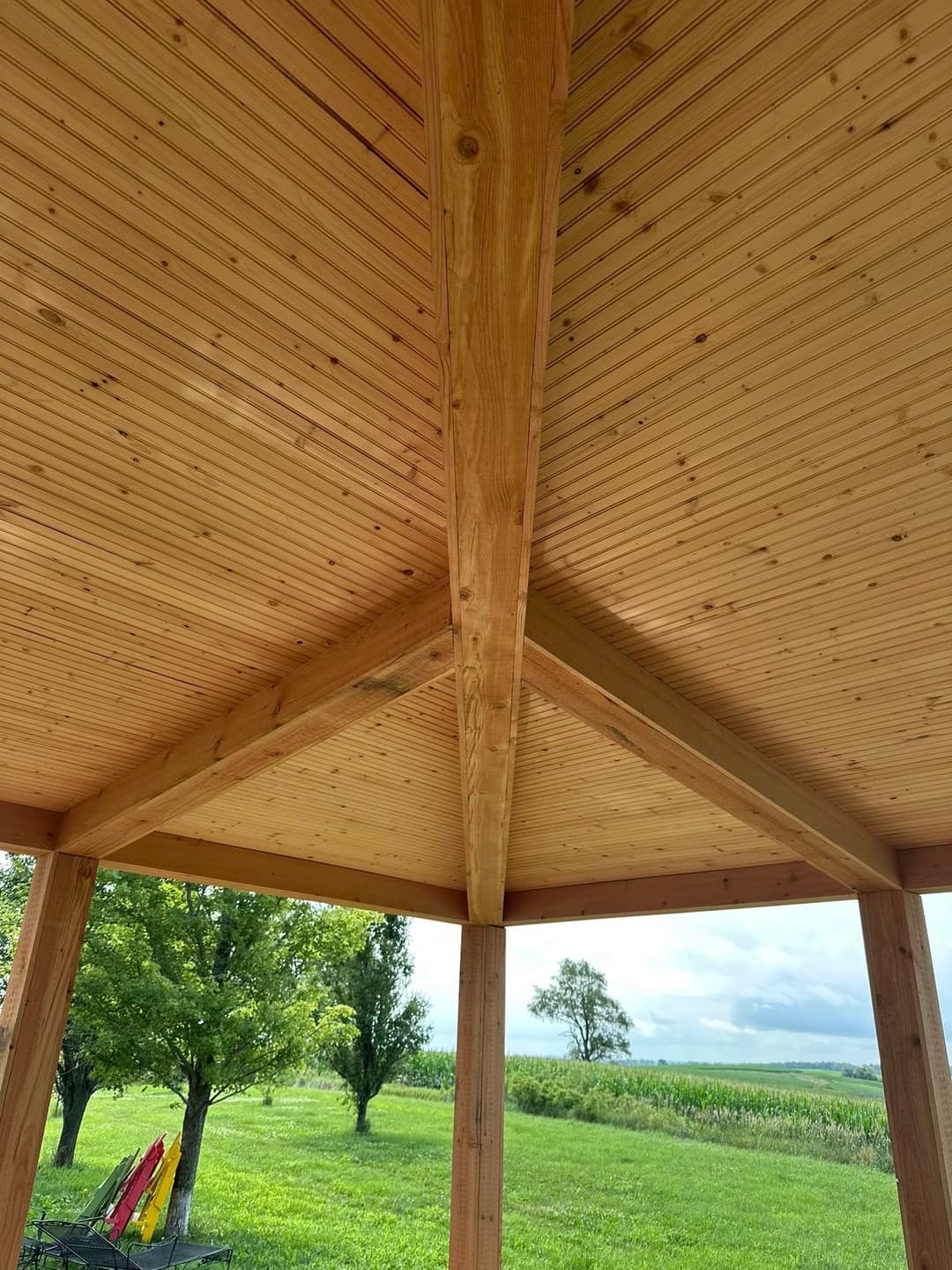 Wooden gazebo ceiling with beam structure, overlooking green fields and trees.