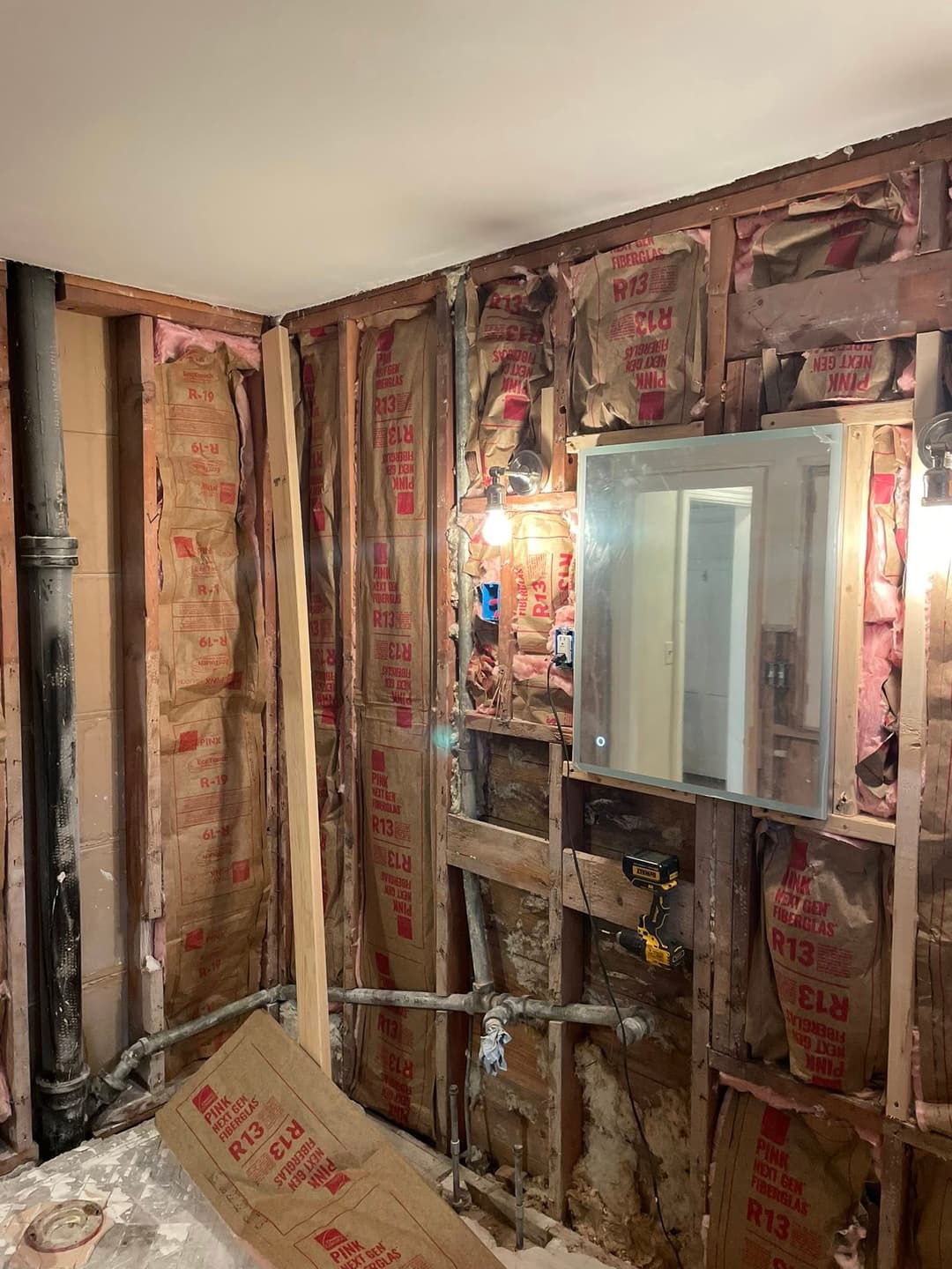 Exposed bathroom wall with insulation, plumbing pipes, and a mirror in a renovation project.