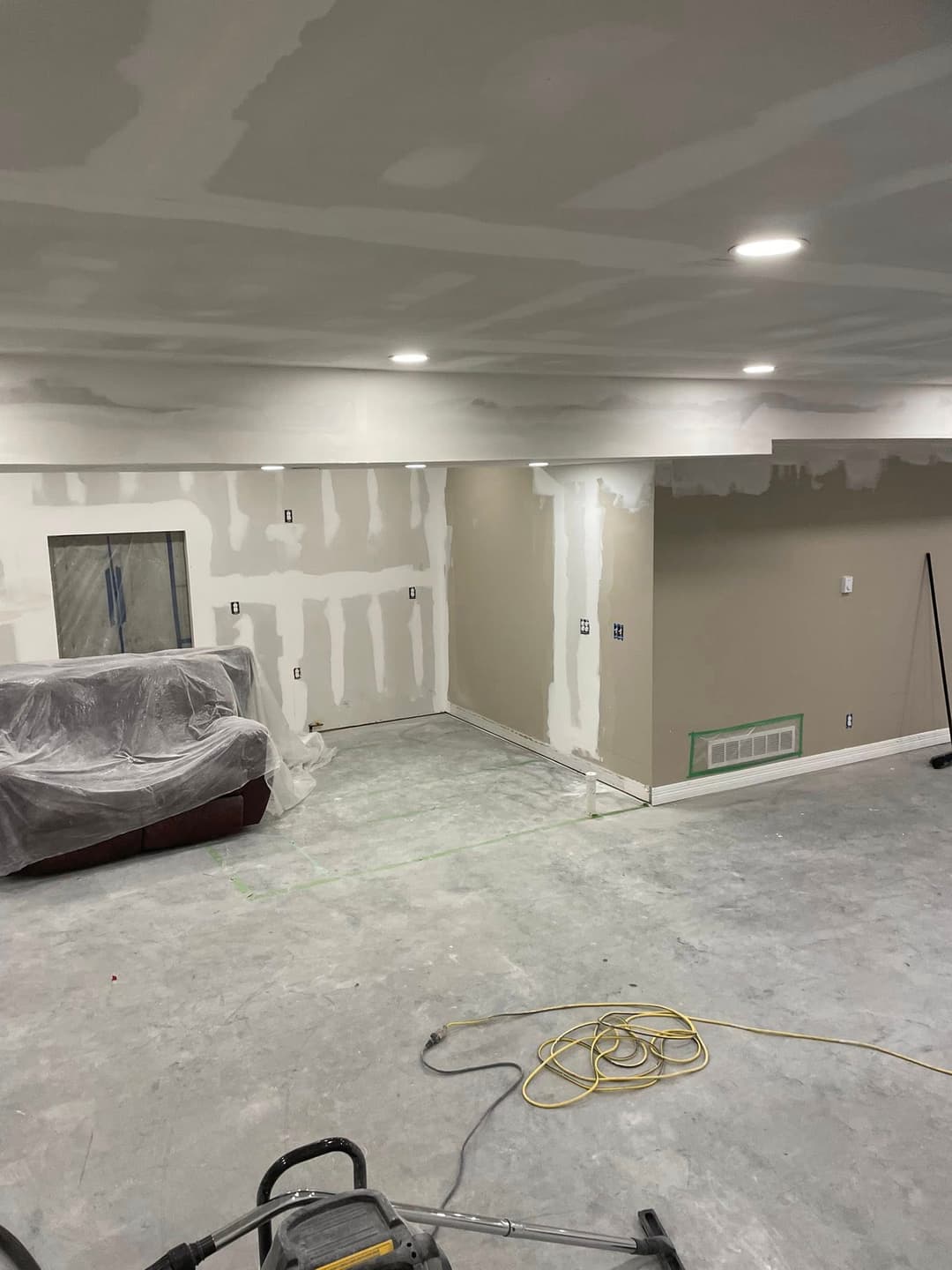 Room renovation in progress with drywall and lighting work, showcasing a couch and construction tools.