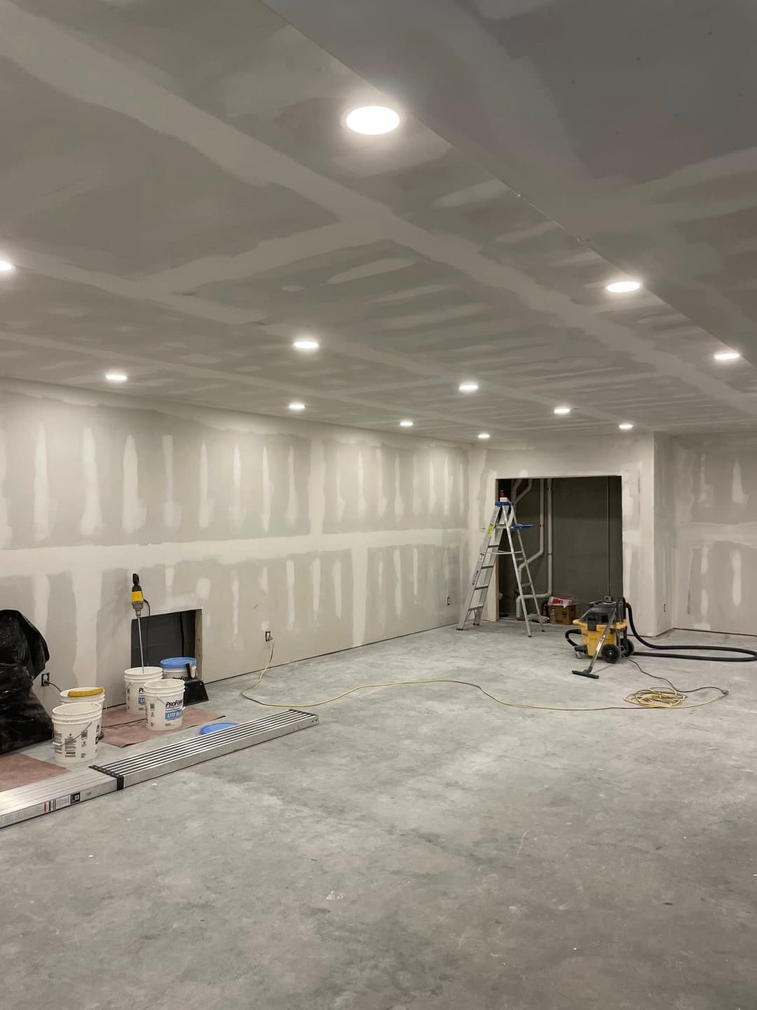 Newly drywalled room with recessed lighting, construction tools, and unfinished flooring.
