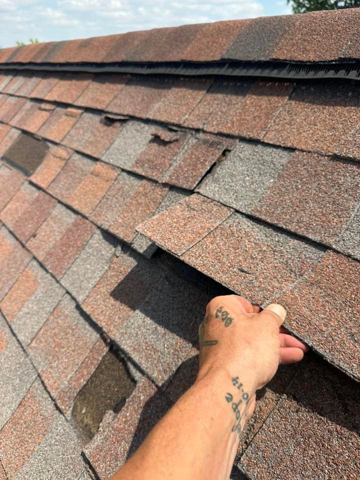 Hand inspecting damaged asphalt shingles on a roof under a clear sky.