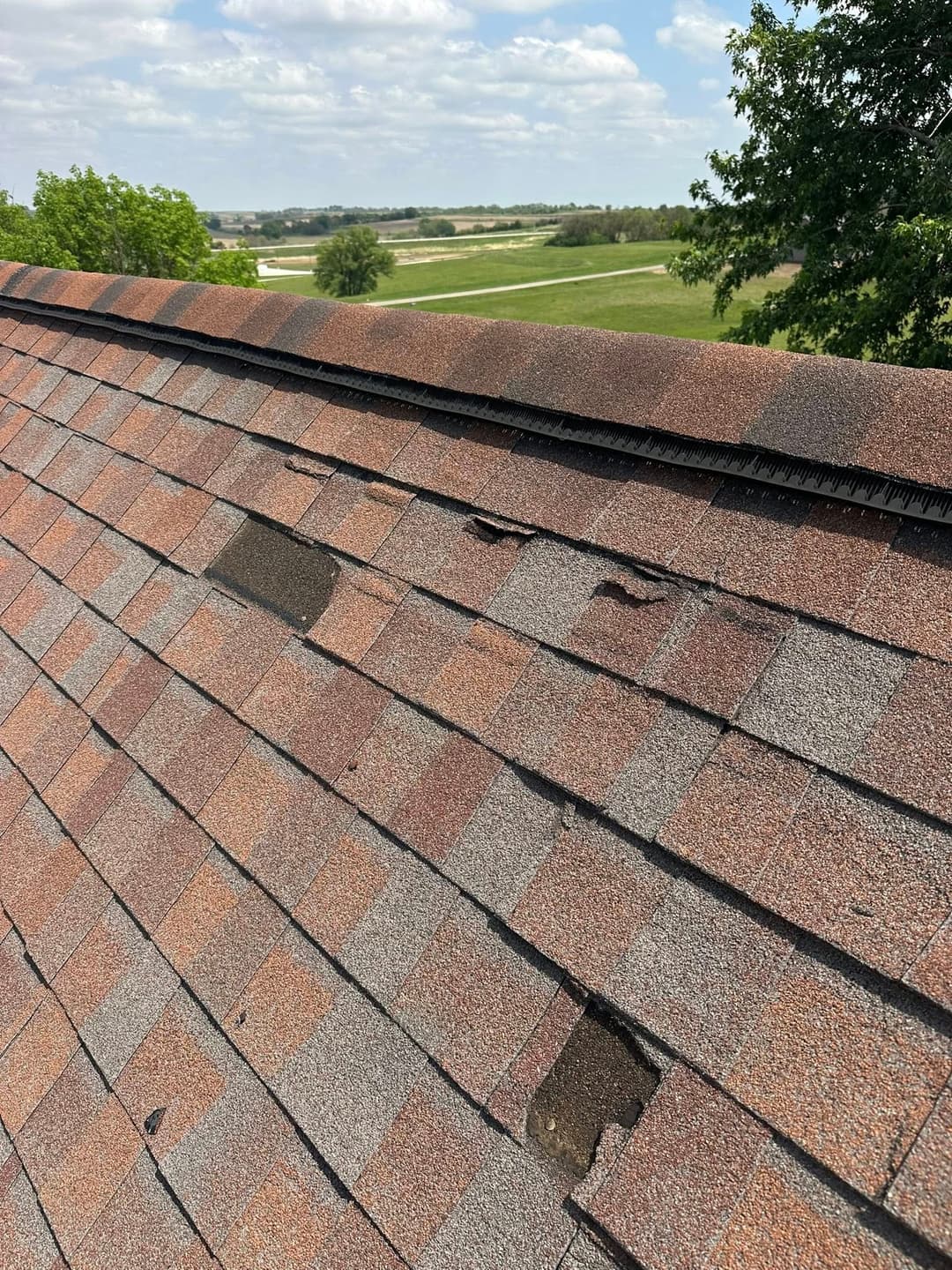 Damaged asphalt shingle roof with missing pieces, showcasing a rural landscape in the background.