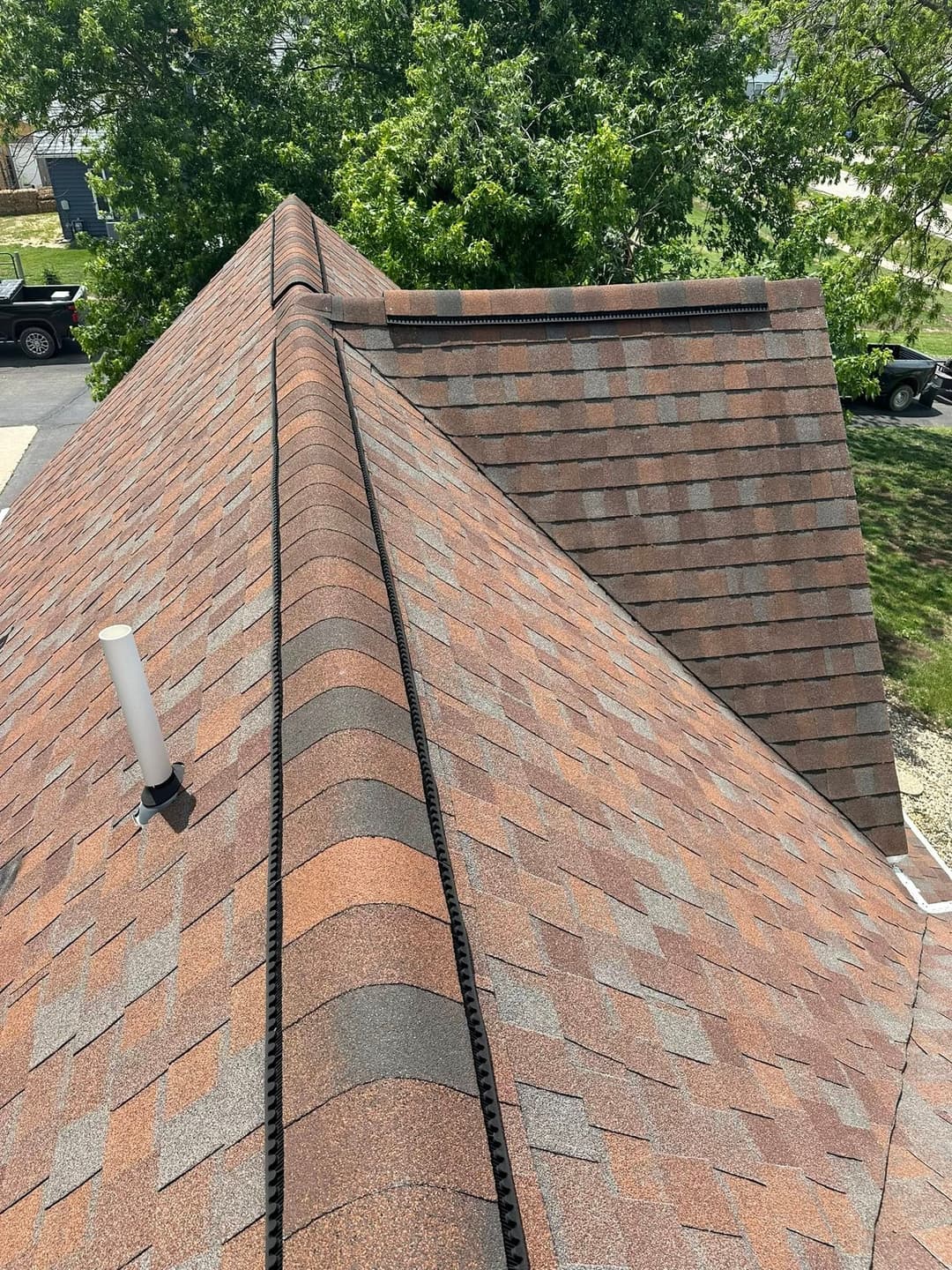 Aerial view of a house roof with red-brown shingles and ventilation pipe against green trees.