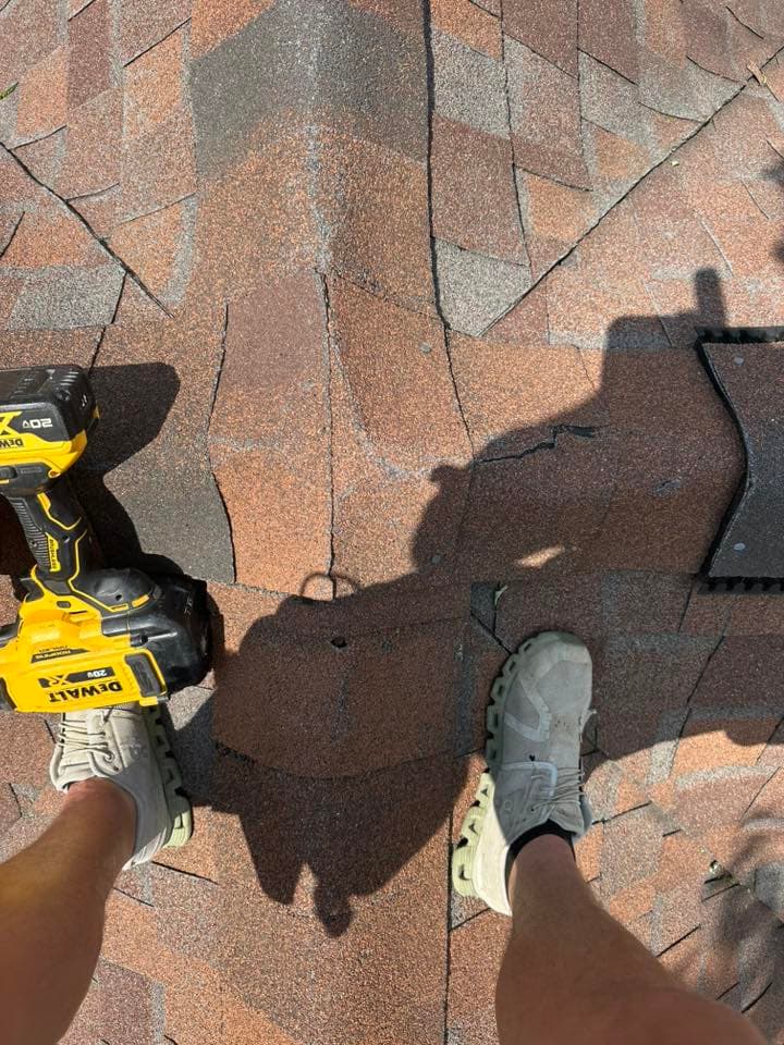 Person's feet on a roof with shingles, holding a power drill, sunlight casting shadows.