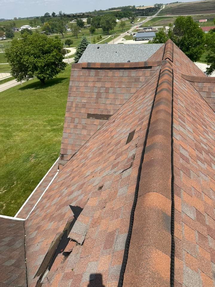 Damaged asphalt shingles on a sloped roof, visible from above, with green landscape in background.