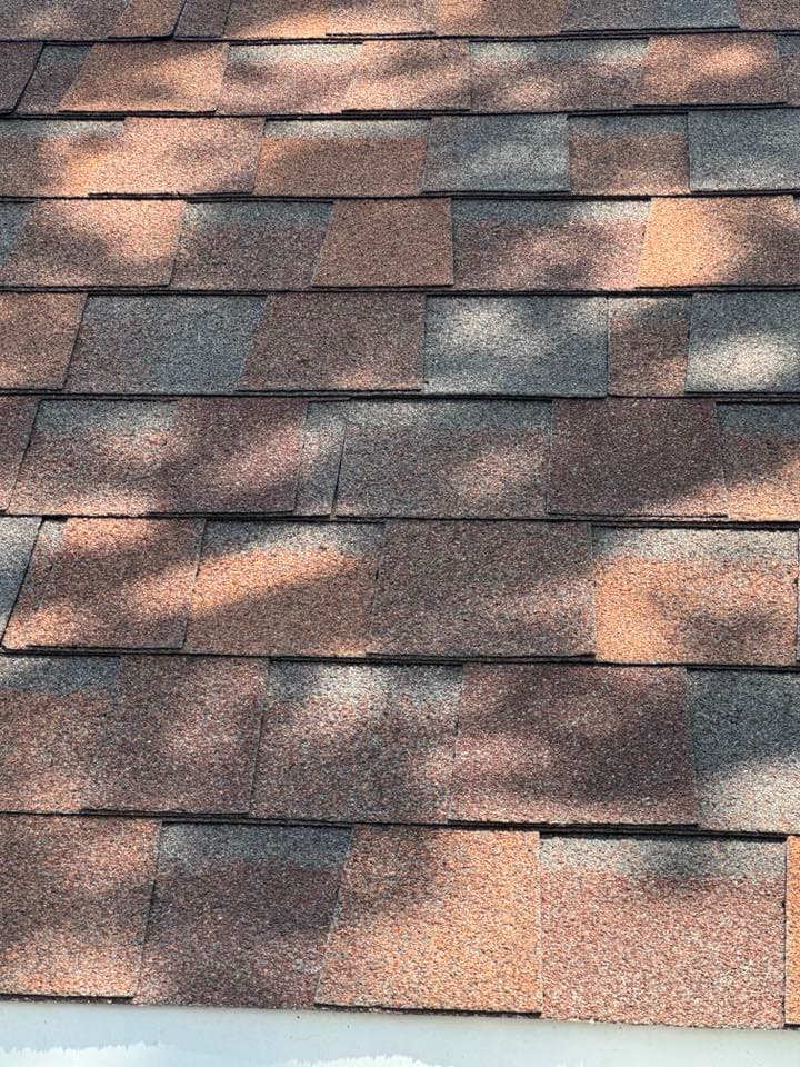 Close-up of asphalt roof shingles in shades of gray and brown with sunlight shadows.